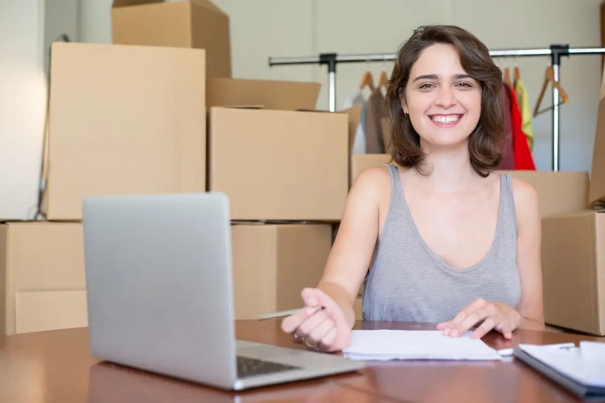 Una emprendedora sonríe frente a su laptop, rodeada de cajas listas para enviar.