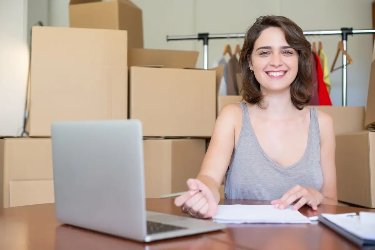 Una emprendedora sonríe frente a su laptop, rodeada de cajas listas para enviar.